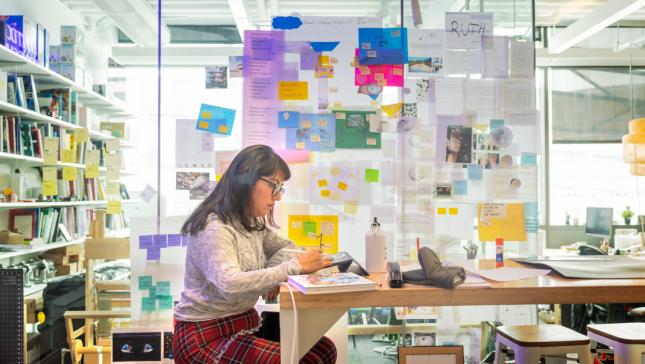 A color photo of a woman working at a desk, surrounded by notes.