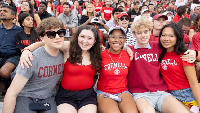 5 students sit front row on the bleechers wearing Cornell red t-shirts with their arms wrapped around one another and smiling