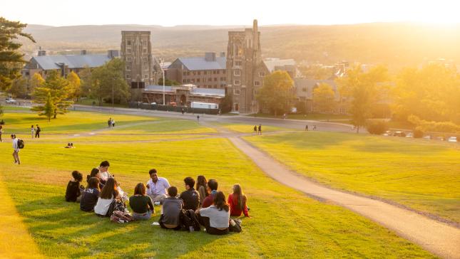 students sit on a light green libe slope during sunset overlooking baker hall