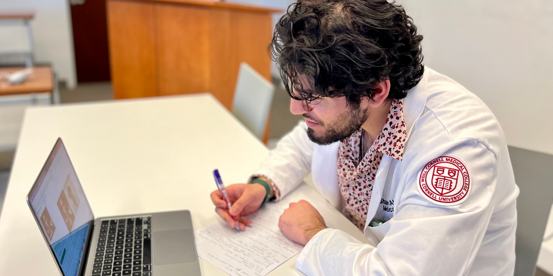 A medical student in a lab coat takes notes in front of a laptop