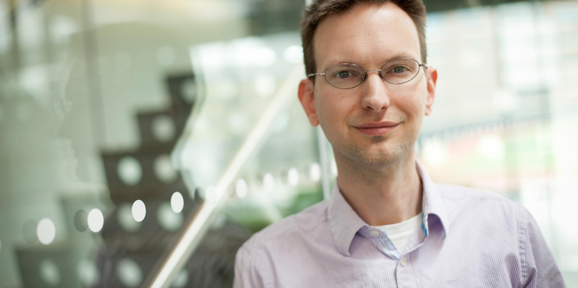 man with short, brown hair and glasses, leans against glass wall