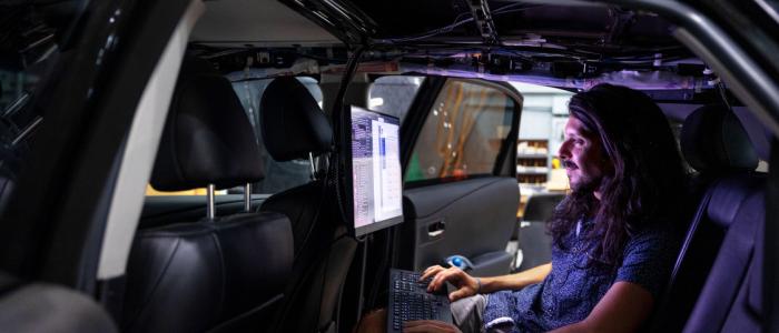 inside of a dark car a man with long dark hair sits in the backset looking at a computer screen