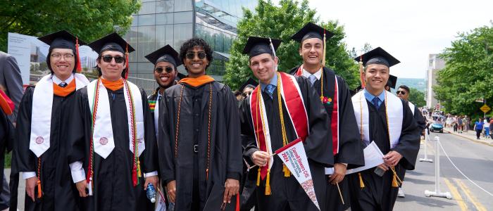 7 male students dressed in black graduation caps and gowns smile in front of Gates Hall