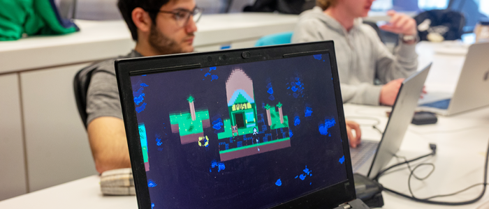 Students working on laptops with a pixel-art game displayed on a screen in the foreground.