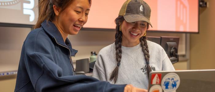 two female students point at a laptop with a red slide in the background