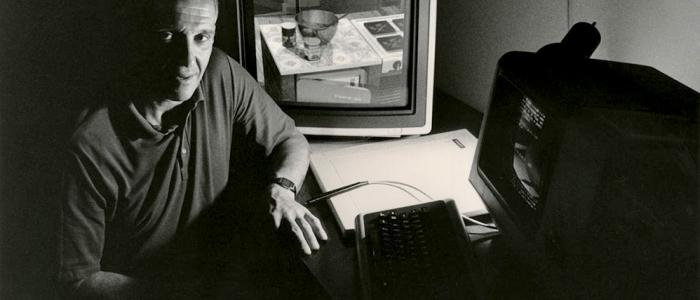 Man seated beside vintage computer monitors displaying early 3D graphics.