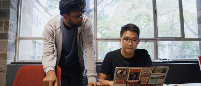 two students (one standing, one sitting) look at computer laptop