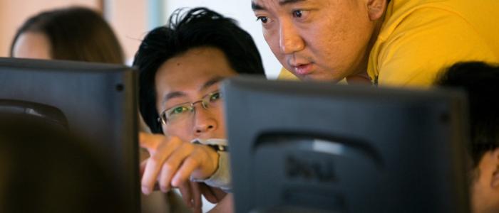a man stands next to another man wearing glasses pointing at something on the desktop computer