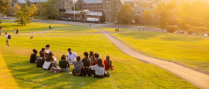 students sit on a light green libe slope during sunset overlooking baker hall