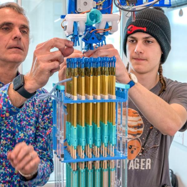 A color photo showing 2 men working on a knitting device.