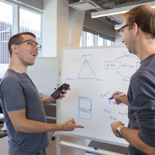 A color photo of 2 men discussing a project in front of a white board.