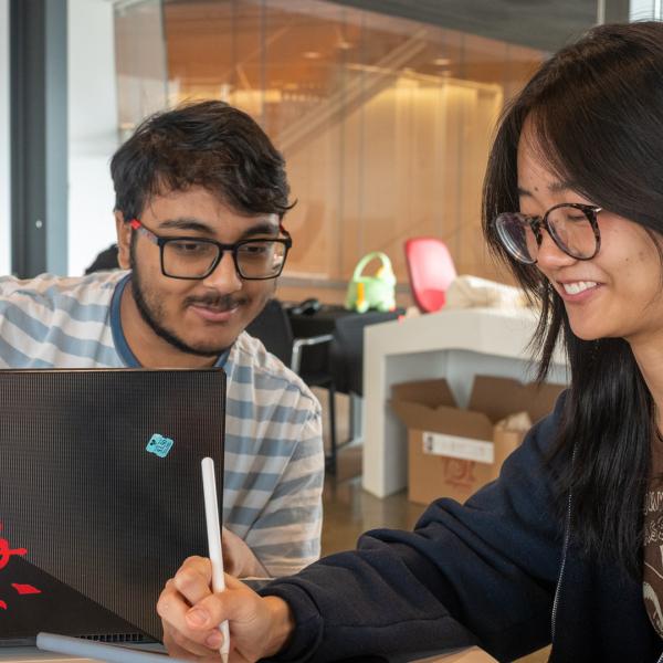Two students sit around a laptop computer with a Flordia license plate bumper sticker. They wear glasses and smile, looking down at a tablet. 