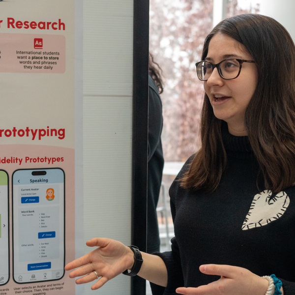 a female student with long brown hair and glasses presents in front of a poster
