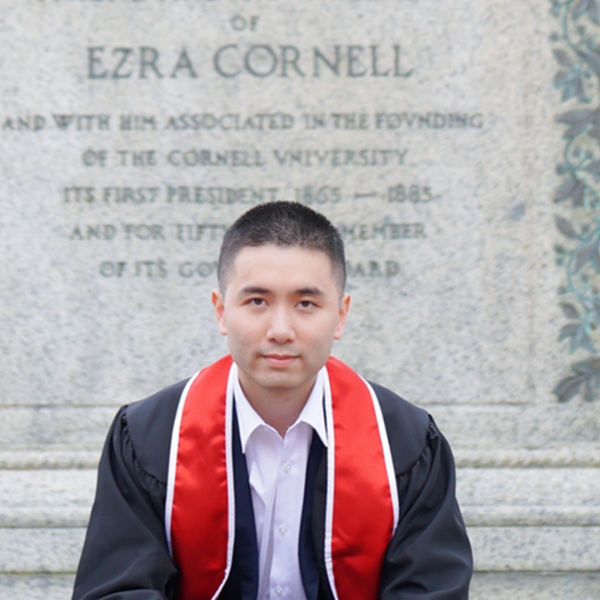 a young asain man sits on a gray statue wearing a black graduation robe and red
