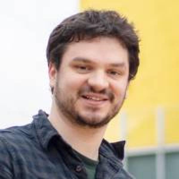 a man with short brown hair and a beard smiles in front of a white and yellow background