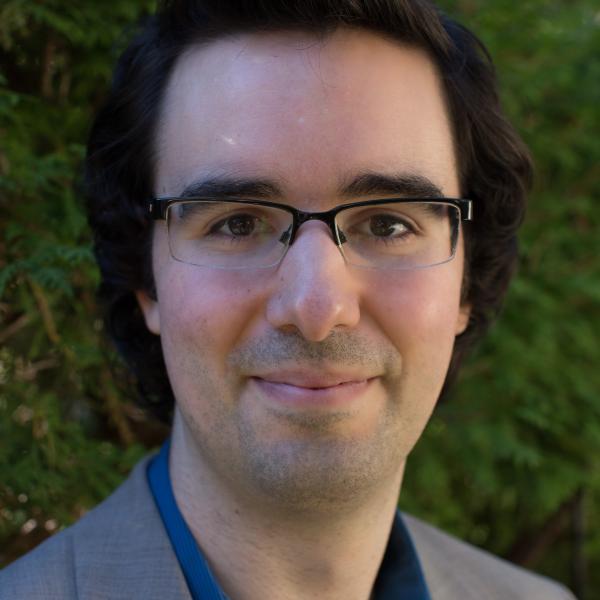 A photo of John Thickstun, a man with short black hair, glasses, and a blue shirt with a gray jacket in front of a leafy background