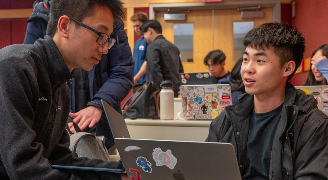 Two students talk across a laptop