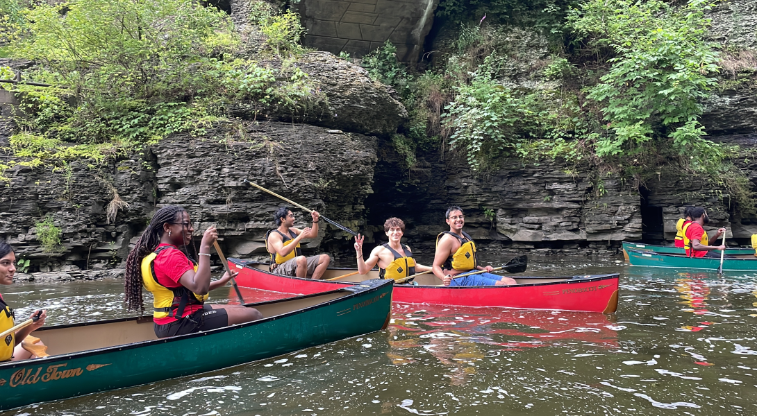 Students wear yellow life vests as they canoe on a lake.