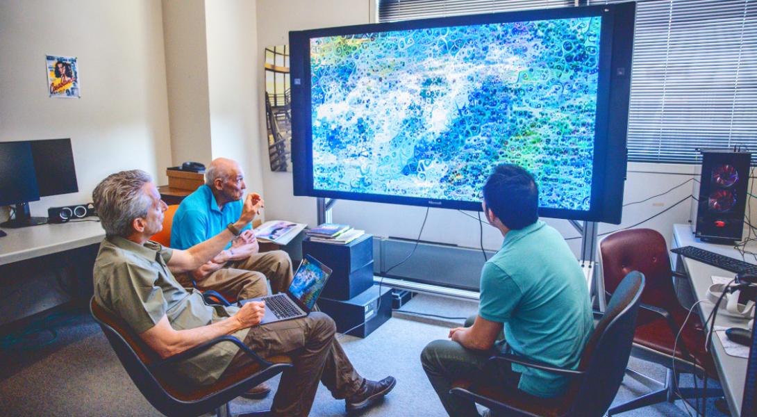 Three people sit in front of a large screen displaying a natural landscape.