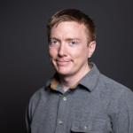 A photo of Adrian Sampson, a man with short dark-blond hair and a gray shirt in front of a gray background