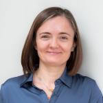 A photo of Alexandra Silva, a smiling woman with shoulder length brown hair and a blue shirt in front of a gray background 