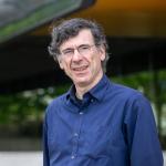 A portrait of Jon Kleinberg, a man with curly gray hair, glasses and a blue shirt, in front of Gates Hall