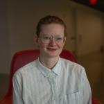 A photo of Sarah Dean, a woman with glasses and blond hair pulled back and a white shirt, sitting in a red chair.