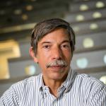 A photo of Fred Schneider, a man with dark gray hair and a mustache, in a striped shirt in front of a staircase