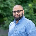 A photo of Sainyam Galhotra, a man with a shaved head, a dark beard, dark glasses and a blue shirt in front of a leafy background.