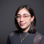 A headshot of Giulia Guidi, a woman with shoulder length brown hair, a gray sweater, brown glasses and gold necklaces.