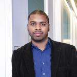 A photo of Hakim Weatherspoon, a mwan with buzzed black hair and beard and mustache, in a blue shirt and black jacket, in a hallway