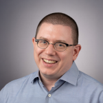 A color photo of Michael Clarkson, a smiling man with a buzzed head, glasses and a gray shirt in front of a gray background