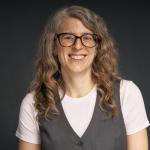A color photo of a woman with long curly brown hair and glasses in front of a gray background
