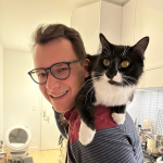 A color photo of a man with glasses smiling for a photo while a black and white cat sits on his shoulders.