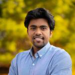A photo of Saikat Dutta, a man with dark brown hair and a beard, wearing a blue shirt in front of a leafy background