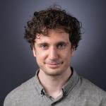 A photo of Kevin Ellis, a man with curly brown hair wearing a beige-gray shirt in front of a gray backround.