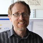 The head and shoulders of a man with glasses, reddish hair and beard, and a striped shirt.