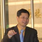 man talking, wearing a dark suit with blue tie and yellow shirt