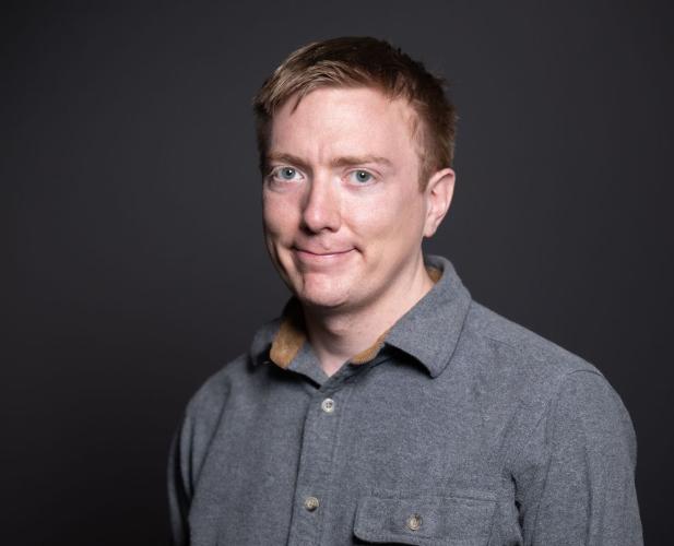 A photo of Adrian Sampson, a man with short dark-blond hair and a gray shirt in front of a gray background