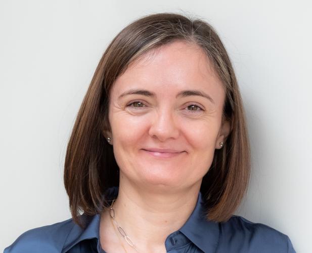 A photo of Alexandra Silva, a smiling woman with shoulder length brown hair and a blue shirt in front of a gray background 