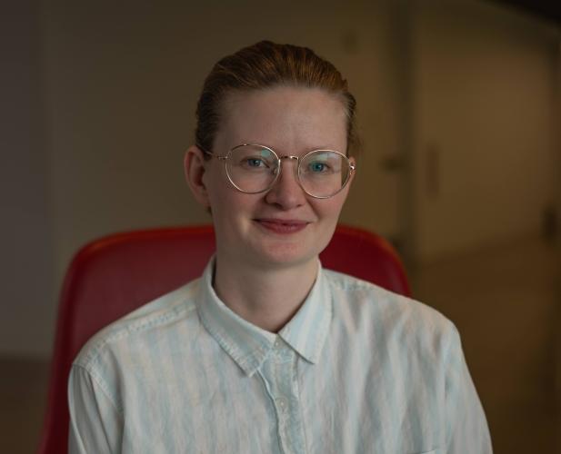 A photo of Sarah Dean, a woman with glasses and blond hair pulled back and a white shirt, sitting in a red chair.