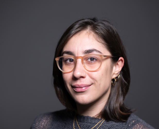 A headshot of Giulia Guidi, a woman with shoulder length brown hair, a gray sweater, brown glasses and gold necklaces.