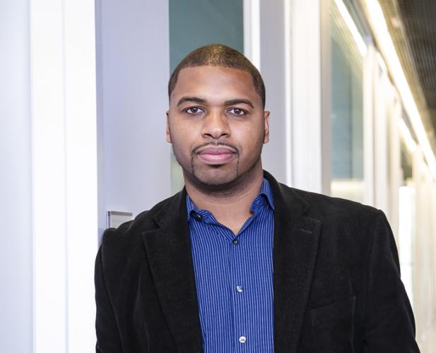 A photo of Hakim Weatherspoon, a mwan with buzzed black hair and beard and mustache, in a blue shirt and black jacket, in a hallway