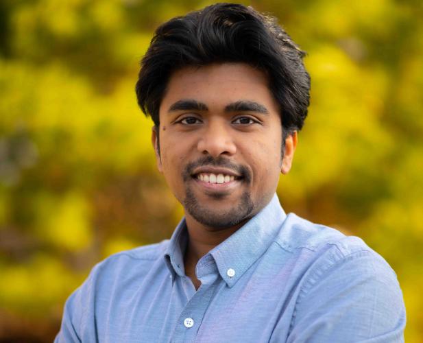A photo of Saikat Dutta, a man with dark brown hair and a beard, wearing a blue shirt in front of a leafy background