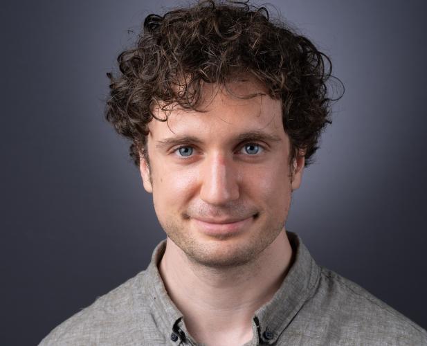 A photo of Kevin Ellis, a man with curly brown hair wearing a beige-gray shirt in front of a gray backround.