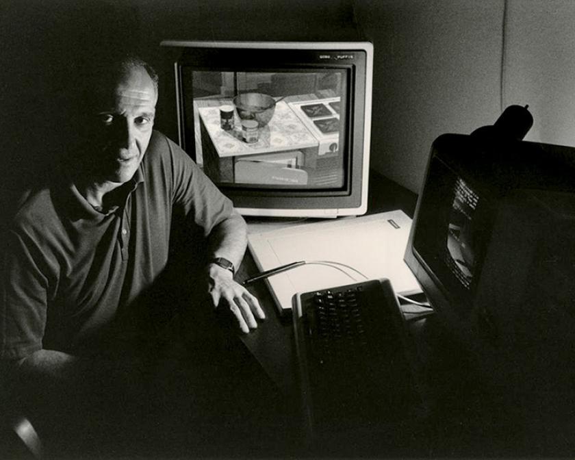 Man seated at a desk with vintage computer monitors displaying early 3D graphics in a dimly lit room.