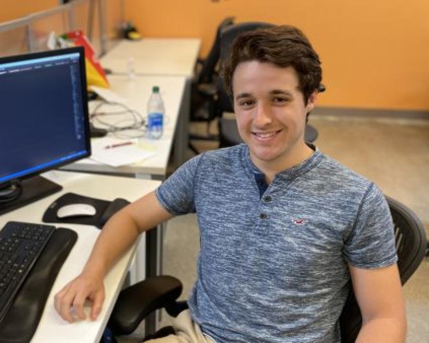 A student smiles and looks at the camera while sitting at a desk with two computer monitors