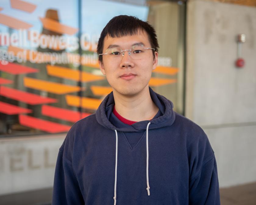 a male asian student with black hair, glasses and wearing a navy pullover hoodie stands in front of red and orange painted windows