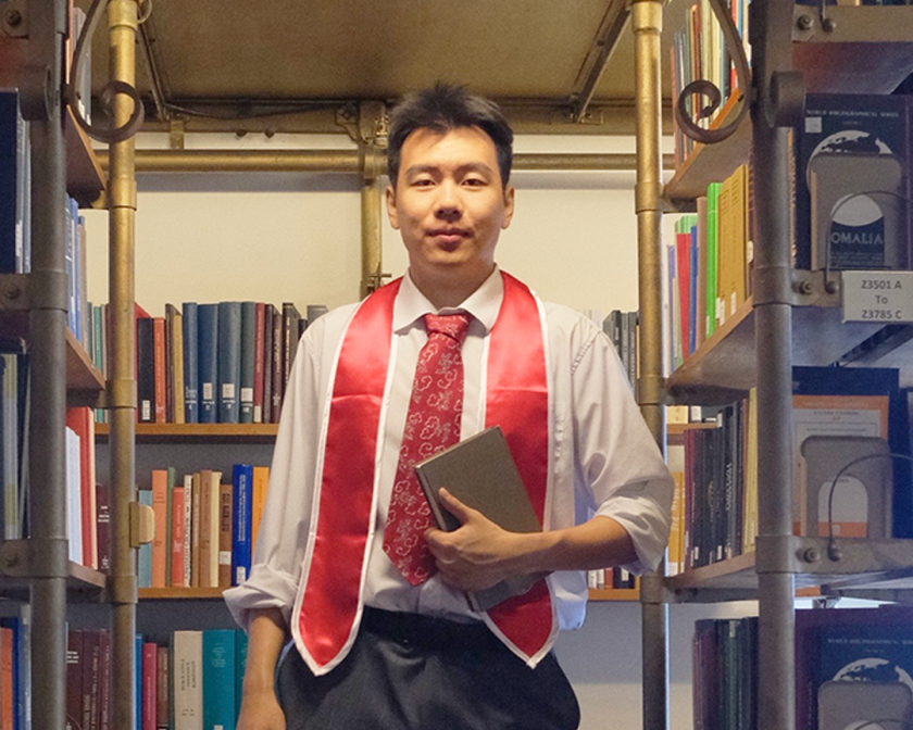 A color photo of a man standing in a library aisle holding a book.