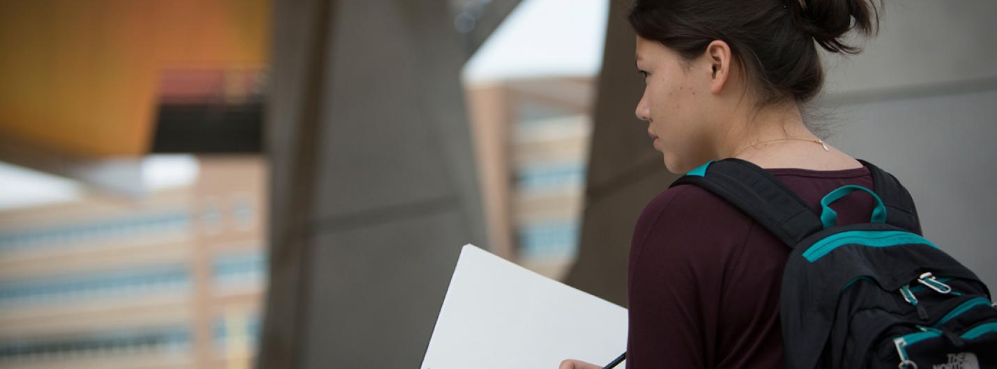 a female student walks towards an orange colored building holding a piece of paper